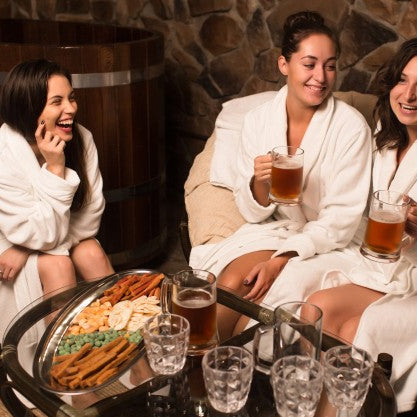 women enjoying cold beer while sitting outside a sauna and wearing a bathrobe 